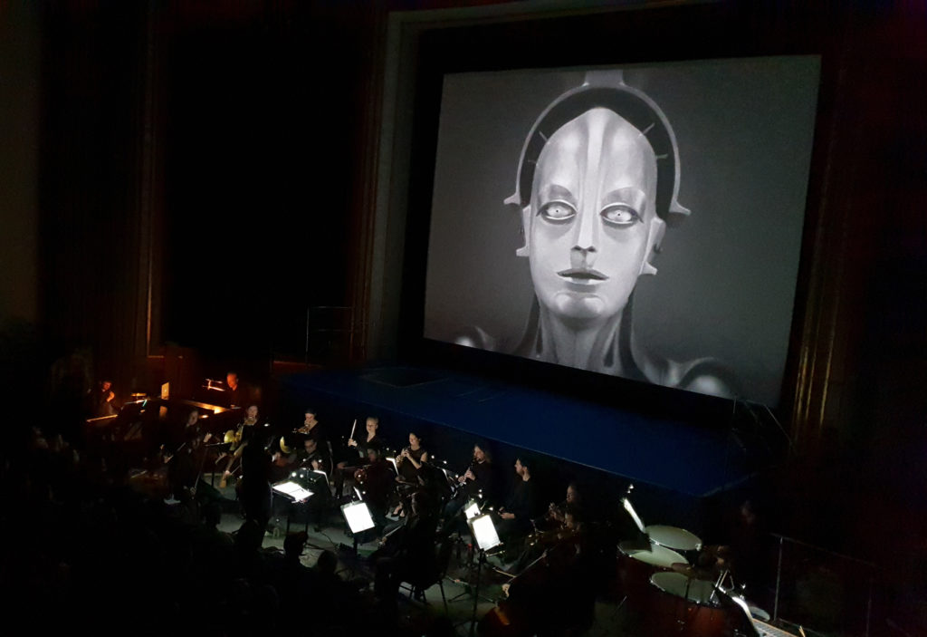 Inside Babylon's cinema. On the screen is a still from Fritz Lang's Metropolis, which is accompanied by a orchestra who play in front of the stage.