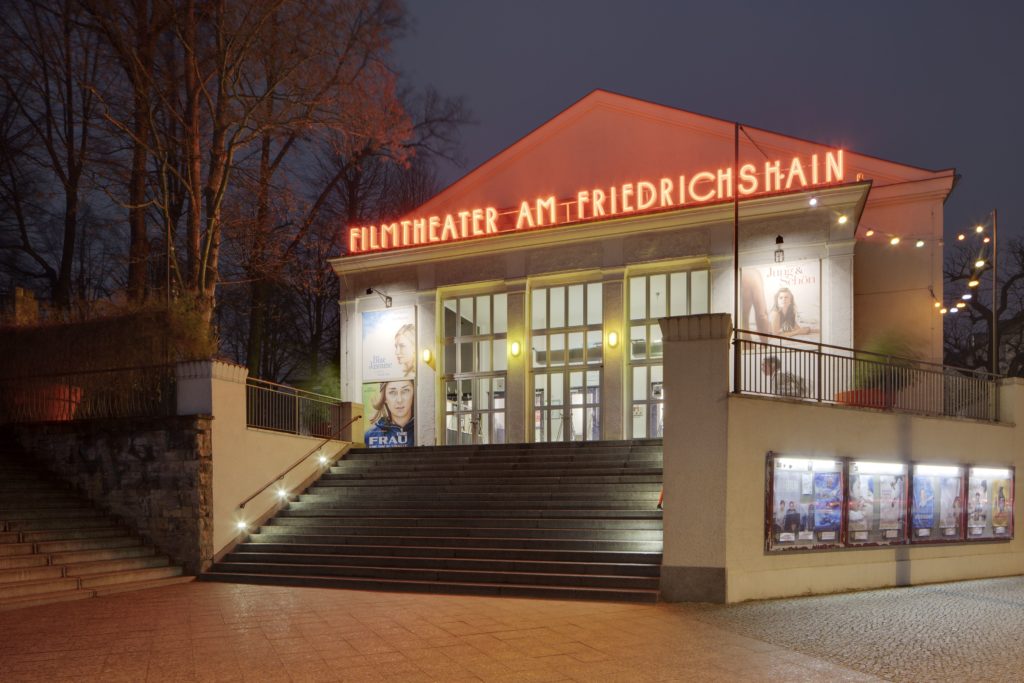 Entrance to Filmtheater am Friedrichshain with a red neon sign that says "Filmtheater am Friedrichshain". On the walls there are film posters.