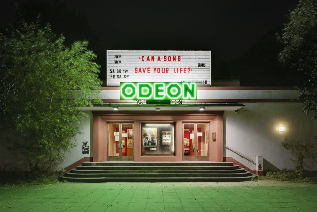 Entrance to the Odeon cinema. Above the green neon sign that says "Odeon" is a white sign showing different screening times.