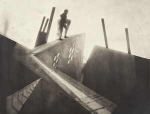 A still image from German expressionist classic The Cabinet of Dr. Caligari. The image depicts wildly distorted buildings where a male figure carries a woman across the roofs of the buildings.