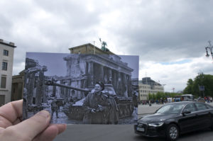 An image by Michael Beaton where he holds an old photograph of the Brandenburg Gate at the end of the war in front of the Brandeburg Gate today. The film image depicts a young soldier in front of a tank and surrounded by rubble.