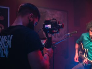 Photo by Jose P. Ortiz that depicts the collaboration between a filmmaker and a guitarist. In the foreground is the filmmaker holding a gimble and in the background the guitarist wears a green tshirt and strums the guitar