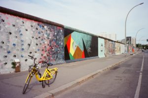 Image of the East Side Gallery section of the Berlin wall. A yellow bike sits in the foreground with the street-art covered wall behind.