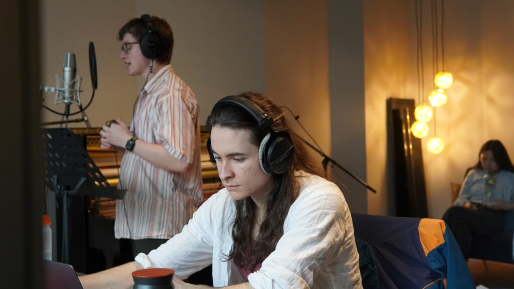 In the foreground, a Music Production student sits at his computer. He is wearing headphones. In the background, a vocalist sings into a microphone.