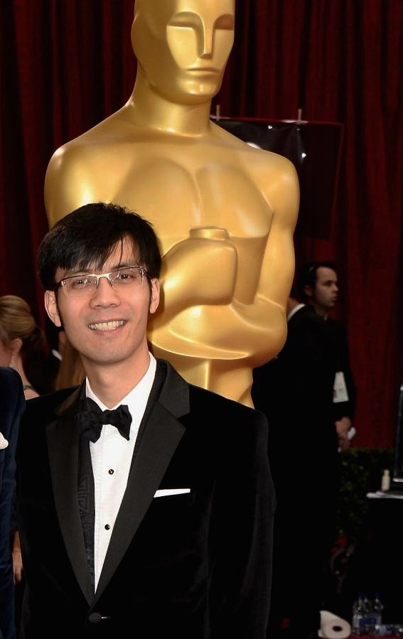 Baldwin Li standing in front of large Oscar statue wearing a tuxedo suit and smiling